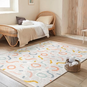May include: A white and gray area rug with a colorful abstract pattern of pink, yellow, and blue squiggles. The rug is placed in a room with a wooden bed and a stuffed animal on the rug.