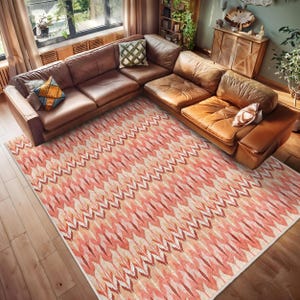 May include: A pink and cream area rug with a geometric pattern. The rug is laid out on a wooden floor in a living room setting with a brown leather sectional sofa.