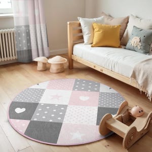 Puede incluir: Una alfombra redonda de patchwork rosa y gris con estrellas y corazones blancos. La alfombra está en una habitación con una cama de madera y almohadas.