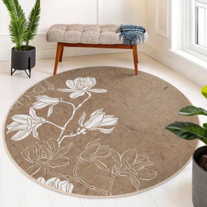May include: Round brown rug with a white magnolia flower design. The rug features a detailed floral pattern with a neutral color palette, suitable for various interior styles. The rug is a decorative floor covering.