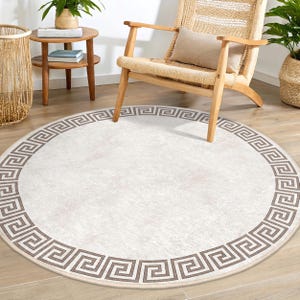 May include: A white round rug with a brown Greek key border. The rug is on a wooden floor and a chair is in the background.