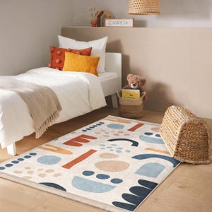 May include: A white bed with a beige throw blanket and orange and yellow pillows. A brown teddy bear sits on a woven basket next to the bed. A colourful abstract rug with blue, orange, and beige shapes is on the floor. The rug has the word "CARPETA" printed on it.