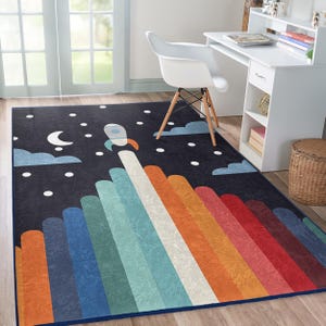 Op de afbeelding: Een kleurrijk tapijt met een raketontwerp op een donkerblauwe achtergrond. De raket lanceert in een hemel vol witte sterren en wolken. Het tapijt heeft een regenboog van kleuren, waaronder rood, oranje, geel, groen, blauw en paars.