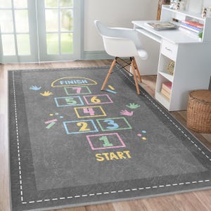 Op de afbeelding: Een grijs speelkleed met een hopscotch-ontwerp met kleurrijke nummers van 1 tot en met 7. Het kleed heeft een gestikte rand en de woorden "START" en "FINISH" zijn op het kleed gedrukt.