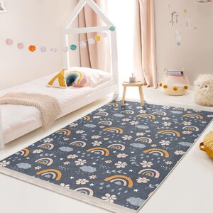 Könnte beinhalten: Ein grauer und weißer Teppich mit Regenbogen-, Wolken- und Blumenmuster. Der Teppich hat einen weißen Fransenrand und liegt in einem Kinderzimmer mit einem weißen Bett und einem Holzhocker.