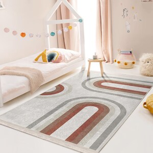 May include: A white and gray area rug with a red, gray, and white geometric pattern of arches. The rug is in a child's bedroom with a white bed and a white and gray throw blanket.