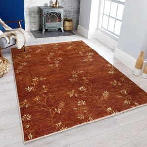 May include: A rectangular rust-colored rug with a cream border. The rug features a repeating pattern of gold-colored branches and leaves. The rug is placed on a light-colored wooden floor in a living room setting.
