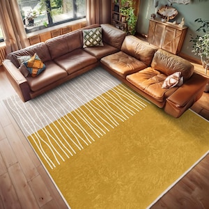 May include: A modern area rug with a white and gray geometric pattern on the top half and a solid mustard yellow on the bottom half. The rug is placed on a hardwood floor in a living room with a brown leather sectional sofa.