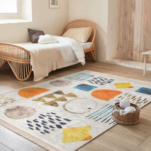 May include: A white and beige area rug with a geometric pattern of orange, yellow, blue, and gray shapes. The rug is on a wooden floor in a room with a wicker bed.