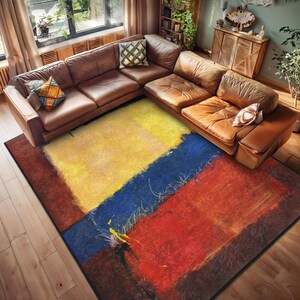 May include: Abstract area rug with a yellow, blue, red, and brown design. The rug is on a wooden floor in a living room with a brown leather sectional sofa.