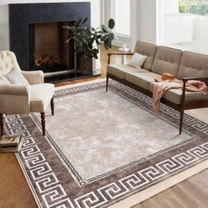 Puede incluir: Una alfombra beige con un borde geométrico marrón y blanco. La alfombra está en un salón con un sofá y una chimenea.