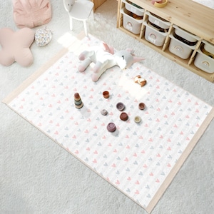 Op de afbeelding: Een witte speelmat met een roze en grijs hartjespatroon. De mat heeft een beige rand en ligt op een wit, pluche tapijt. Er liggen verschillende houten speelgoedjes op de mat, waaronder een stapel ringen en kleine ronde stukjes.