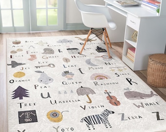 Op de afbeelding: Een wit alfabetkleed met kleurrijke illustraties van dieren, voorwerpen en letters. Het kleed heeft een dinosaurus, een spook, een huis, een sleutel, een straaljager, een blad, een net, een sinaasappel, een peer, een koningin, een regenboog, een boom, een paraplu, een viool, een walvis, een röntgenfoto, een jojo en een zebra.