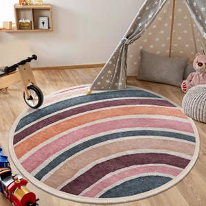 May include: A round, multi-colored rug with a striped pattern in shades of pink, orange, beige, and blue. The rug has a white border and is laid out on a wooden floor.