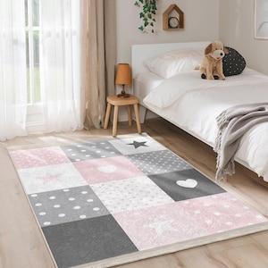 Puede incluir: Una alfombra de patchwork rosa y gris con un diseño de estrellas, corazones y lunares. La alfombra está en un dormitorio con una cama blanca y un taburete de madera.
