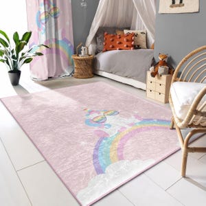 Può includere: Un tappeto rosa e bianco con un unicorno che indossa occhiali da sole e sta in piedi su un arcobaleno. Il tappeto è in una stanza con un letto a baldacchino e una sedia in vimini.