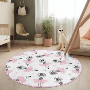 May include: A round white rug with a pink border features a repeating pattern of ballerina girls in pink tutus. The rug is on a light wood floor in a room with a white bed, a chair, and a plant.