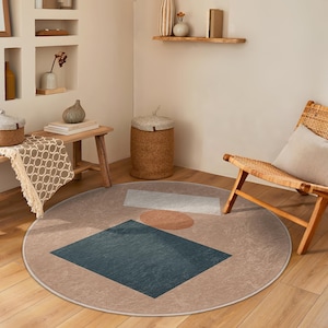 May include: A round, beige rug with a geometric design featuring a blue square, a white rectangle, and a brown circle. The rug is in a living room setting with a wooden chair and a woven basket.