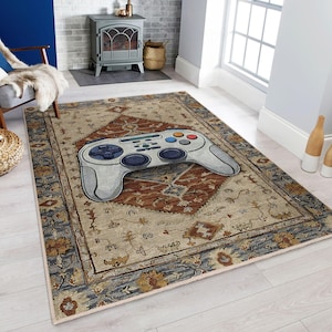 Puede incluir: Una alfombra rectangular con un diseño grande de mando de videojuego de estilo caricatura. La alfombra tiene un fondo beige con un patrón central de diamante en óxido y azul, y un borde detallado. El mando es gris claro con botones azules, naranjas y verdes.