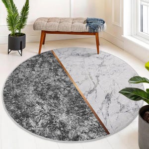 May include: Round area rug with a modern design. The rug is divided diagonally, with one half featuring a dark gray, textured pattern and the other half showcasing a white marble-like design. A thin, copper-colored line separates the two sections.