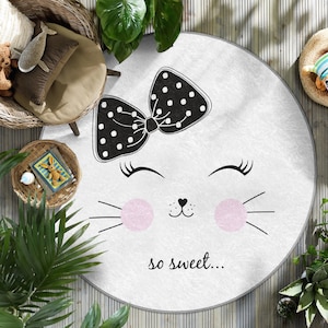 Puede incluir: Una alfombra redonda blanca con un diseño de cara de gato blanco y negro. El gato tiene un lazo negro con lunares blancos en la cabeza. El texto "so sweet..." está impreso debajo de la cara del gato.