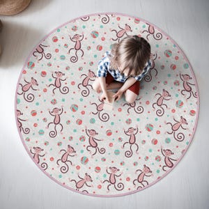 May include: A round pink play mat with a white background and a repeating pattern of monkeys and balls. The monkeys are pink and have brown eyes and black noses. The balls are red, pink, and blue.