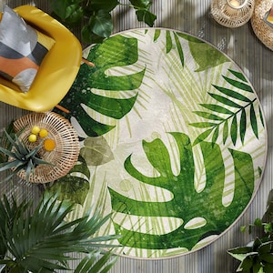 Puede incluir: Una alfombra redonda con un vibrante estampado de hojas tropicales verdes sobre un fondo crema. El diseño de la alfombra incluye grandes hojas de monstera y otro follaje tropical. La alfombra está rodeada por un borde beige claro. Está colocada sobre una terraza de madera.