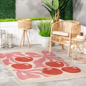 May include: A pink and red cherry patterned area rug with a beige background. The rug is laid out on a patio with a wicker chair, a side table, and potted plants.