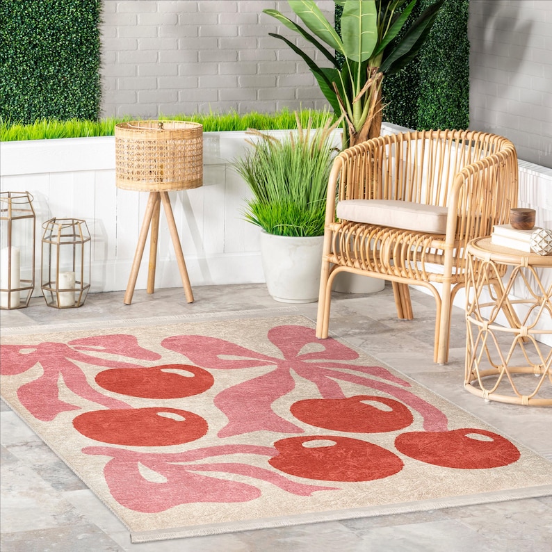 May include: A pink and red cherry patterned area rug with a beige background. The rug is laid out on a patio with a wicker chair, a side table, and potted plants.