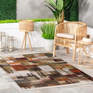 May include: Rectangular outdoor rug with a patchwork design. The rug features a mix of colors including brown, orange, gray, and white, arranged in a geometric pattern. The rug is placed on a concrete patio, with wicker furniture and plants in the background.