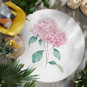 Puede incluir: Una alfombra redonda blanca con una pintura de acuarela de una flor de hortensia rosa con hojas verdes.