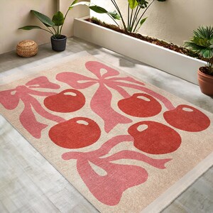 May include: A pink and cream area rug with a pattern of cherries and bows. The cherries are red and the bows are pink.