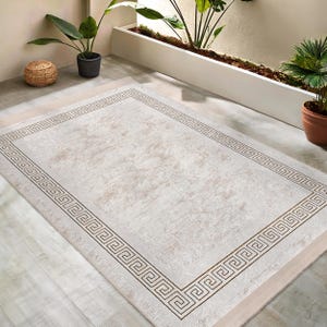 Pode incluir: Um tapete retangular branco sujo com uma borda grega em um tom bege mais escuro. O tapete tem um padrão texturizado sutil e é colocado em um piso claro. Plantas em vasos são visíveis ao fundo.