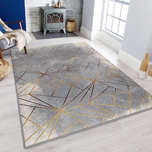 Könnte beinhalten: Ein rechteckiger grauer Teppich mit einem geometrischen Design aus goldenen und braunen Linien. Der Teppich hat ein weiches, strukturiertes Aussehen und liegt auf einem hellen Holzboden. Das Design zeigt sich kreuzende Linien, die sternförmige Muster bilden.