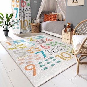 Può includere: Un tappetino da gioco bianco con numeri colorati da 1 a 10, con illustrazioni di vari animali, tra cui pinguini, pesci, granchi e tartarughe. Il tappetino è su un pavimento in legno chiaro in una stanza con un letto a baldacchino bianco e una sedia in vimini.