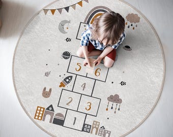 Puede incluir: Una alfombra de juego redonda, gris y blanca con un diseño de hopscotch con los números del 1 al 6. La alfombra también tiene un arcoíris, nubes, estrellas y un cohete. Un niño está jugando en la alfombra.