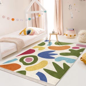 May include: A colourful abstract rug with a white background and various shapes in shades of blue, green, yellow, orange, and red. The rug is laid out on a light wood floor in a room with a white bed and a wooden stool.