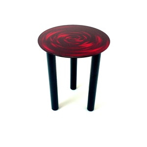 May include: A small, round side table with a striking design. The tabletop features a swirling red pattern, while the legs are black. The table is approximately 18 inches tall and 12 inches in diameter, suitable for various decorative uses.