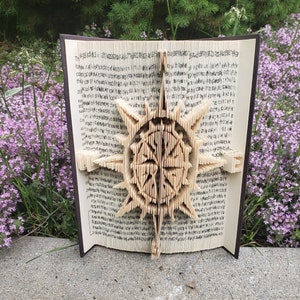 May include: A book with pages folded into a 3D design of a starburst. The book is open and the starburst is in the center of the image. The book is sitting on a concrete surface with green grass and purple flowers in the background.