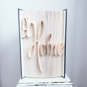 May include: A book art sculpture of the word "Home" created by folding the pages of a book. The sculpture is displayed on a white pedestal.