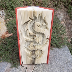 May include: A book with a carved rose design. The pages are cut and folded to create a three-dimensional rose. The book has a red cover and is displayed on a stone surface with greenery in the background.
