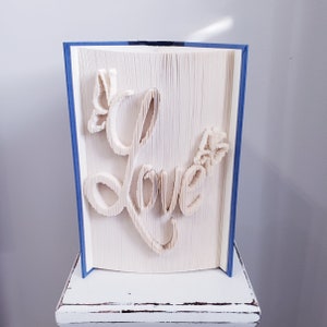 May include: A white book with the word "Love" folded into the pages. The book has a blue cover and is sitting on a white surface.