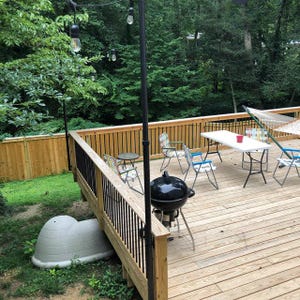 May include: Outdoor deck with a black grill, a white table, and colorful chairs. A hammock is strung up in the background. String lights hang overhead, and a wooden fence surrounds the deck. The scene is set in a wooded area.