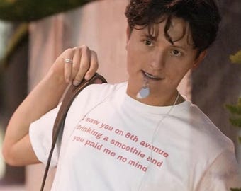 Puede incluir: Una camiseta blanca de manga corta con texto en rojo que dice: I saw you on 8th avenue drinking a smoothie and you paid me no mind. La prenda se lleva con una correa marrón sobre el hombro y un anillo plateado en el dedo.