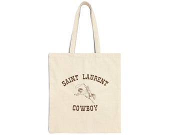 May include: A beige tote bag with brown text that reads "SAINT LAURENT COWBOY". A drawing of a cowboy riding a horse is centered on the bag.