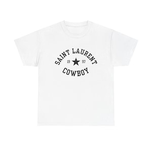 Könnte beinhalten: Weißes T-Shirt mit schwarzem Grafikdesign. Das Design zeigt die Worte "SAINT LAURENT COWBOY" in einem bogenförmigen Layout, mit "19" und "97" auf beiden Seiten eines Sterns.