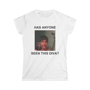 May include: A white short-sleeved t-shirt featuring a centered rectangular graphic print. The print shows a person holding a glass of red wine in a shower setting, with the black text HAS ANYONE SEEN THIS DIVA? printed above and below the image.