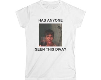 May include: A white short-sleeved t-shirt featuring a centered rectangular graphic print. The print shows a person holding a glass of red wine in a shower setting, with the black text HAS ANYONE SEEN THIS DIVA? printed above and below the image.