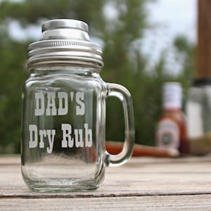 Peut inclure: Un pot en verre transparent avec un couvercle argenté et une poignée. Le pot est étiqueté "DAD'S Dry Rub".