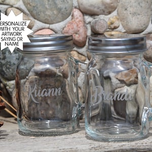 May include: Two clear glass mason jars with silver lids and handles. The jars are personalized with the names "Kiann" and "Brandi".